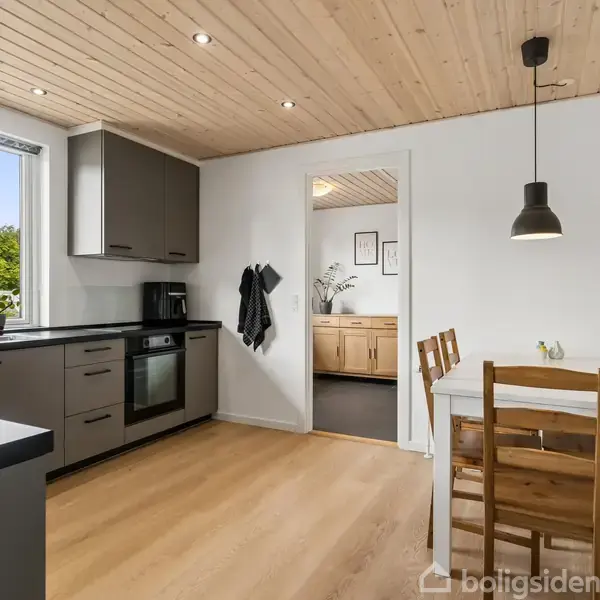 Køkkenbord med blomster står ved et moderne, gråt køkken med komfur og ovn, lyst trægulv og træloft, spisebord med to stole; lys hænger over bordet.