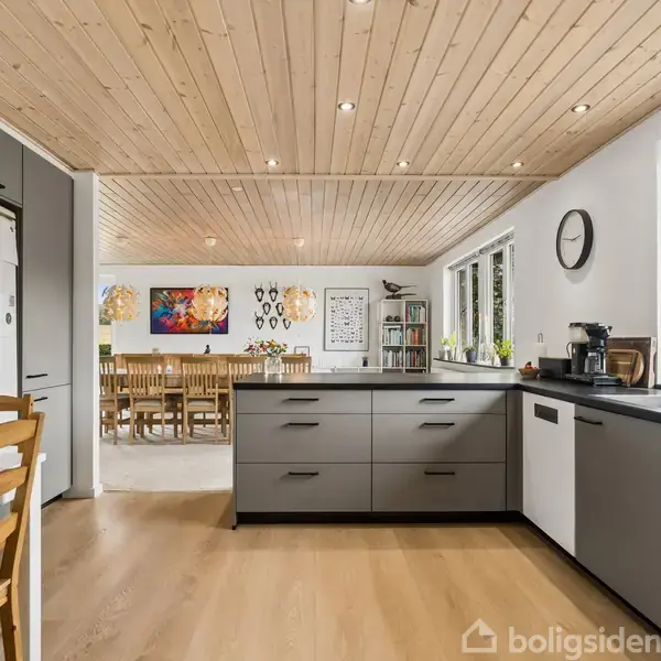 Køkken med grå skabe og sort bordplade, placeret ved store vinduer. Et spiseområde med træbord og stole ses i baggrunden. En tegning hænger på køleskabet. En rund ur hænger på væggen.