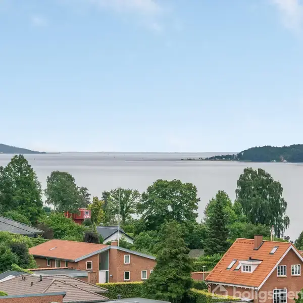 Røde teglhuse står stille ved kysten omgivet af grønne træer. I baggrunden ses en rolig vandudstrækning med bakkede skovklædte områder under en klar blå himmel.
