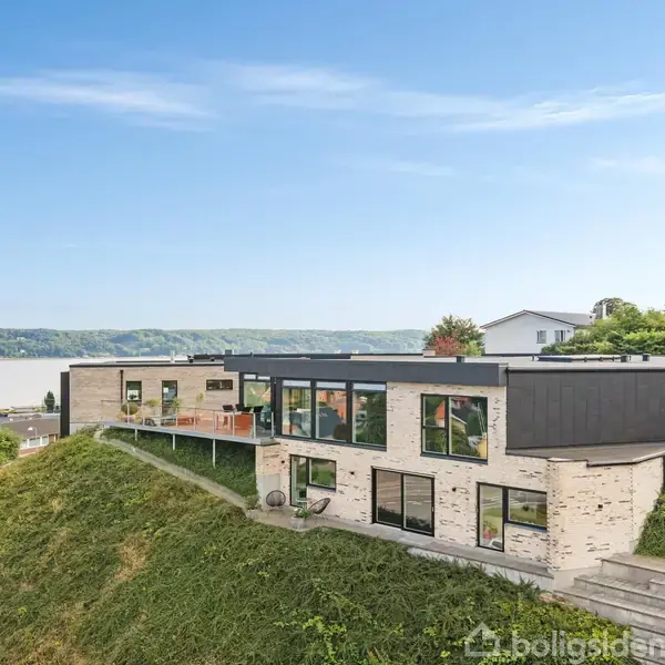 Et moderne, fladt taghus med store vinduer står på en skrånende græsplæne. Huset er omgivet af grønne planter og har udsigt over en bred flod og skovklædte bakker i baggrunden.