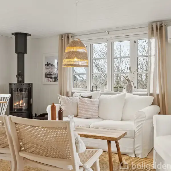 En hvid sofa står i et lyst, hyggeligt stue med en brændeovn ved siden af. Et træbord står foran sofaen, omgivet af en afslappende atmosfære med flere vinduer.