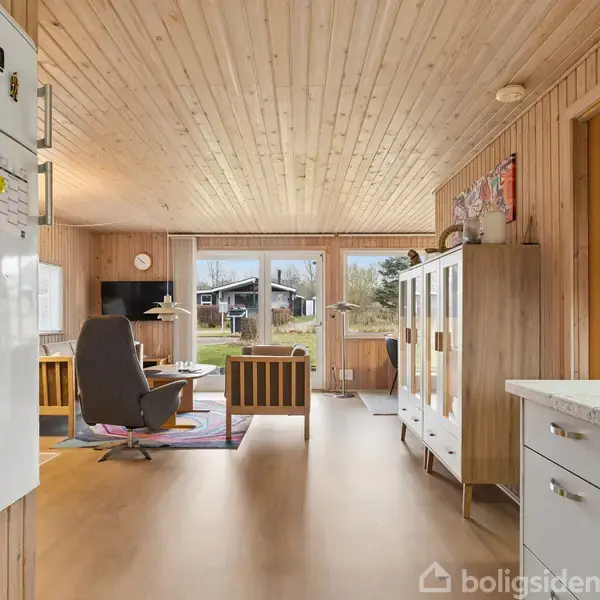 Stue med trævægge og loft, hvor en grå lænestol er placeret ved et sofabord. Store vinduer giver udsigt til en have. Køleskab og skab er i forgrunden.