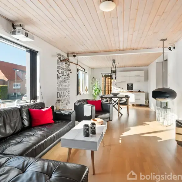 Sort lædersofa med røde puder står i en moderne og lys stue. Ved siden af er et sofabord med dekorative genstande. I baggrunden ses et spisekøkken med træloft og træmøbler.