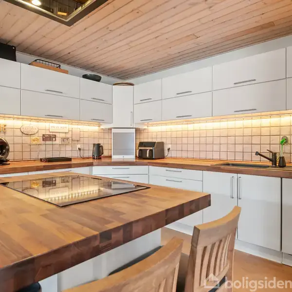 Et moderne køkken med hvide skabe og træbordplade, der omkranser en indbygget kogeplade. En kaffemaskine og brødrister står på bordet. Flisebagvæg og træloft skaber en varm atmosfære.