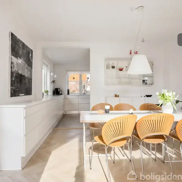 Spisebord med seks stole står i et lyst køkken-alrum. En hylde med hvid service og dekorative planter omkranser rummet. Store vinduer slipper sollys ind og skaber en indbydende atmosfære.