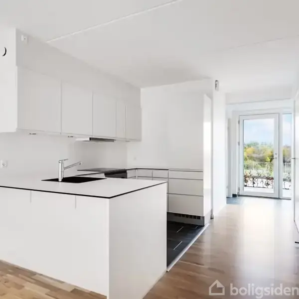 Et moderne køkken med hvide elementer, sort bordplade og indbygget vask. Køkkenet ligger i et lyst rum med trægulv, store vinduer og udsigt til natur. Tekst: "Ribo-ejendomme".