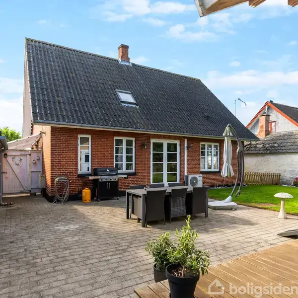 Et rødt murstenshus med sort skråtag, i en gårdhave omgivet af hvide mure med en grey udendørs pavillon, havemøbler og planter i krukker på flisebelagt terrasse.