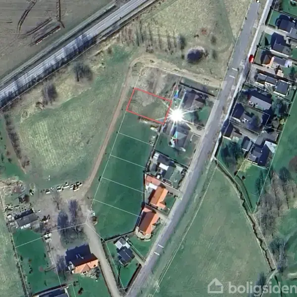 Luftfoto af et boligområde viser en tom byggegrund markeret med en rød kant i centrum. Omkring grunden er der huse, marker og veje.