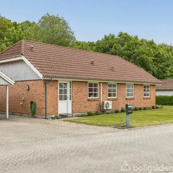 Rødstenshus med brunt tegltag står på en grund med græsplæne og indkørsel. En bil er parkeret i en carport ved siden af huset. Skoven danner baggrund.
