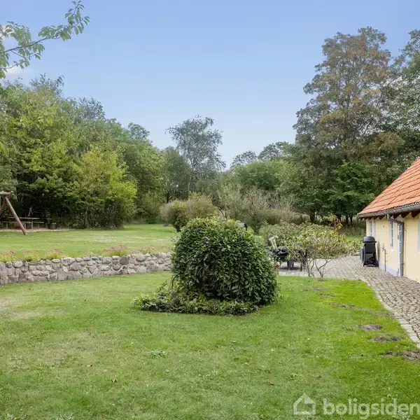 Gult landehus med rødt tegltag står stille i en frodig have omgivet af grønne træer og en græsplæne. Der er træmøbler foran huset midt i det landlige miljø.