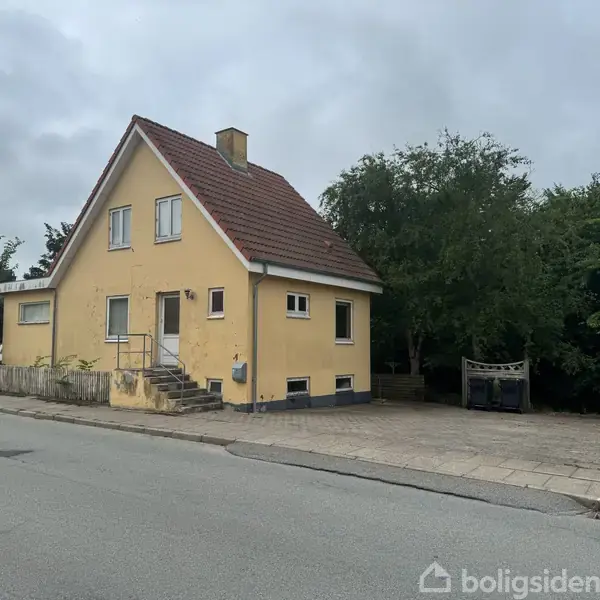 Gul villa med rødt tag står langs en stille vej. Foran huset er der en flisebelagt sti med en træbænk omgivet af grønne buske og træer i baggrunden.
