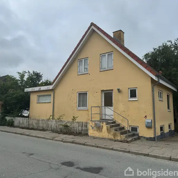 Gulmalet hus med rødt tegltag står på fortovet langs en vej. En trappe fører op til hoveddøren. Huset omgives af buske og træer i et beboelsesområde.
