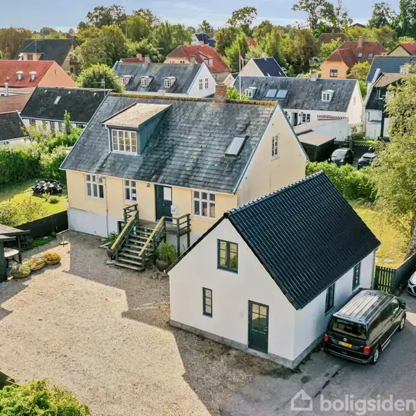 Et stort, gulmalet hus med trappe foran og grå skifertag står i et parcelhuskvarter. Ved siden af er et mindre, hvidt hus og parkerede biler langs vejen.