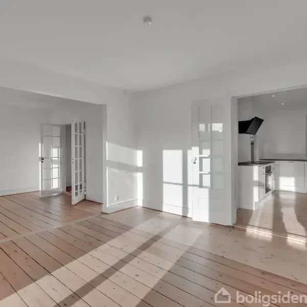 Et lyst rum med trægulve og hvide vægge. To sæt franske døre åbner til et andet rum og et køkken med hvide skabe. Rummet er solbelyst.