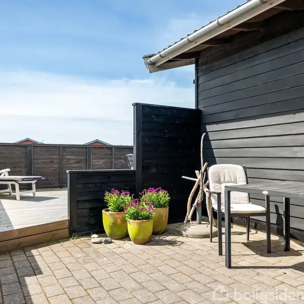 Udendørs terrasse med solstole på et hævet trædæk. Et bord og en stol står foran et sort træhus, omgivet af blomster i farverige krukker. Klar himmel i baggrunden.