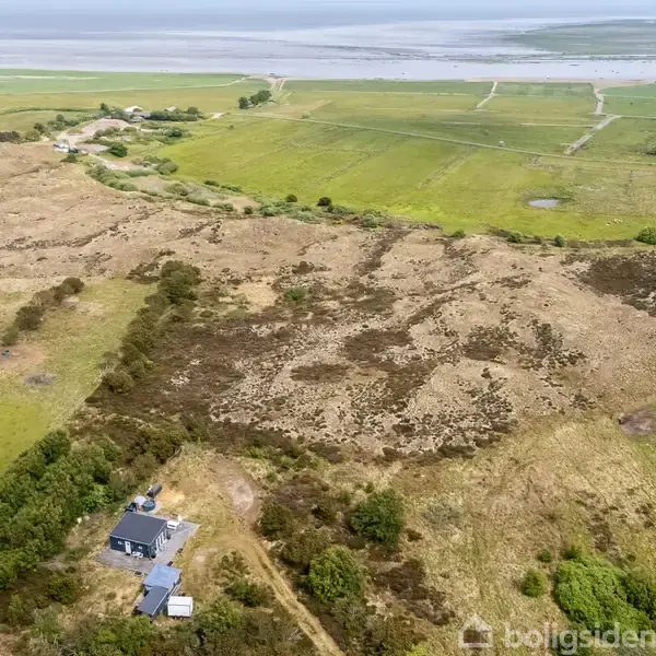 Et hus ligger i bunden af et grønt og bakket landskab. Det omgivende område består af marker og kystlinje i baggrunden, med spredt beplantning og åbne områder.