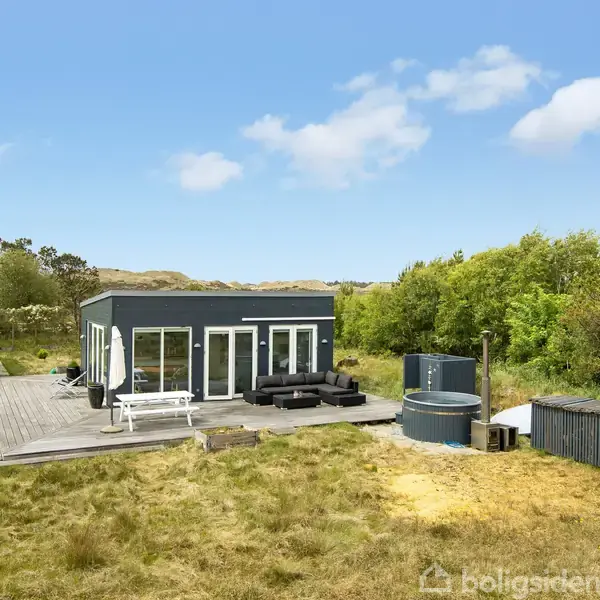 Et moderne sommerhus står på en træterrasse med havemøbler. En lille tilbygning ses ved siden af. Omgivet af græs og træer under en blå himmel. Skilt tekst: "POST BOX".