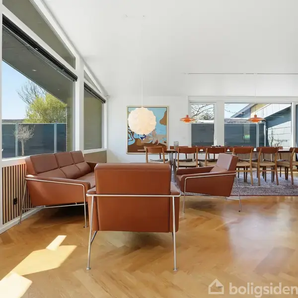 Brune lædersofaer og stole står i en moderne stue med store vinduer, masser af naturligt lys og en udsigt til haven. En spisebordslampe hænger over et langt spisebord.
