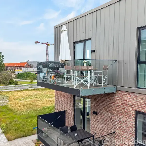 Altan med glasrækværk indeholder et bord og stole med en stor parasol. Den hænger udenfor en moderne bygning med en grå facade. I baggrunden ses en konstruktionskran.