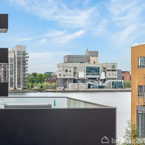 En moderne bygning ligger langs en flod. Bygningen har store glaspartier og geometriske former. I baggrunden ses flere bygninger og en blå himmel med lette skyer.