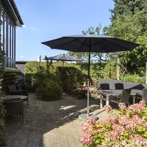 En terrasse med bord og stole under en stor parasol. Omgivet af tætte buske, blomsterbede og et højt træ. Sollys skinner gennem havemøblernes skygger, hvilket skaber en hyggelig atmosfære.