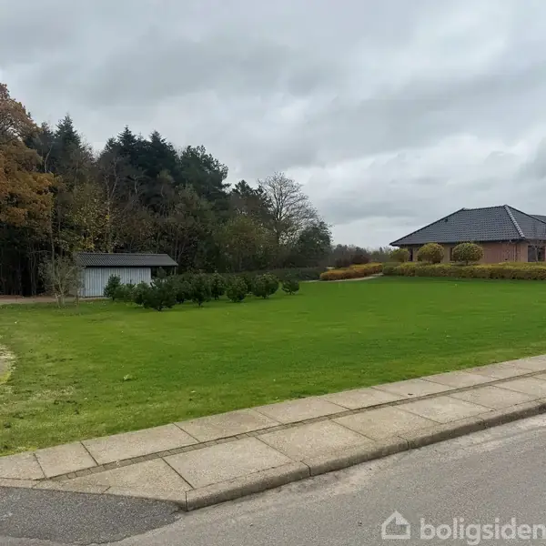 En grøn græsplæne grænser op til en flisebelagt sti, omgivet af buske og træer. Til højre ses et moderne hus og til venstre en mindre bygning.