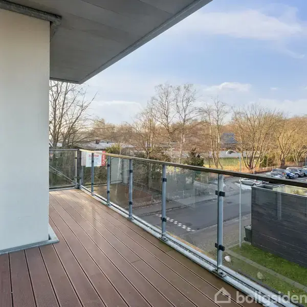 En åben altan med glasrækværk, der fører ud til en udsigt over træer og en parkeringsplads, set fra en moderne bygning med trægulv på altanen.