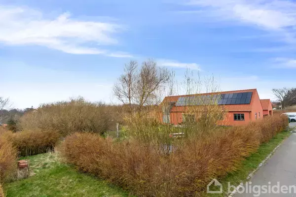 Orange hus med solpaneler på taget, omgivet af tæt buskads og græs. Beliggende ved siden af en vej under en blå himmel med lette skyer.
