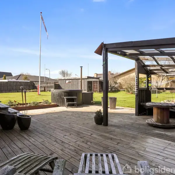 Træterrasse med loungemøbler og plantekrukker, omgivet af en have. I baggrunden ses et flag på stang ved siden af en græsplæne og en afskærmet pavillon.