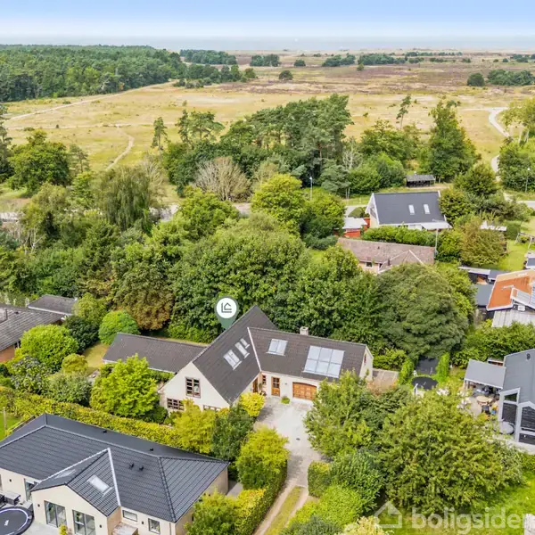 Hus med solpaneler på taget omgivet af grønt i et villakvarter. Omkringliggende huse og en stor, skovrig naturpark i baggrunden.