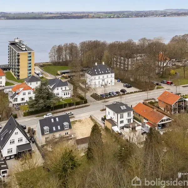 Flere villaer med røde og sorte tage står stille i en række langs en vej. I baggrunden ses en stor bygning og en sø omgivet af træer.