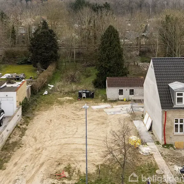 Tomt jordareal ligger mellem to huse med en grusbelagt overflade. En lille bygning står i baggrunden omgivet af træer. Til venstre holder en båd ved siden af et hus.