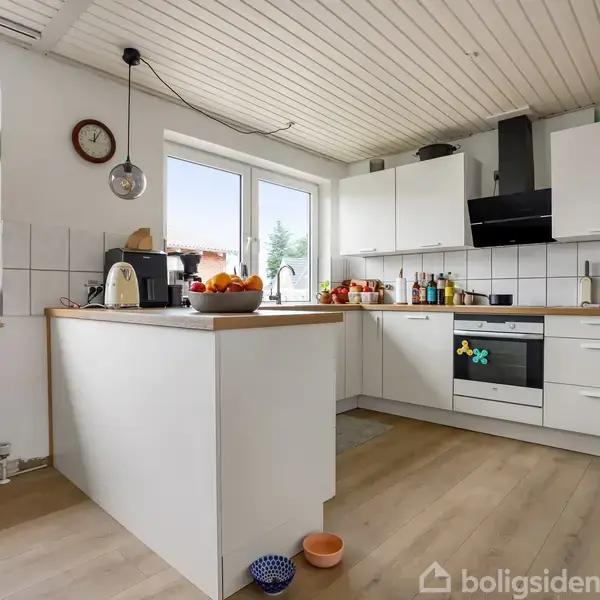 Et moderne køkken med hvide skabe. En kogeplade og en indbygget ovn er synlige. Der står frugtskåle og køkkenmaskiner på bordet, omgivet af store vinduer.