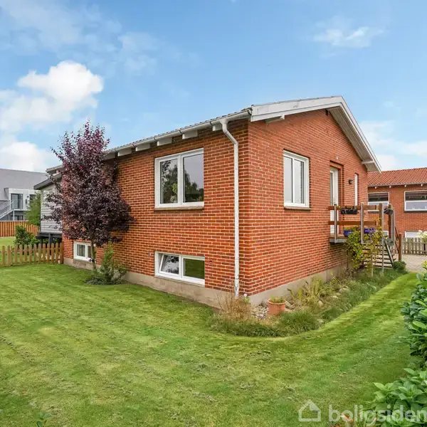 En rød murstensvilla med hvide vinduer, omgivet af en velplejet græsplæne og træer; et hegn omgiver ejendommen i et boligkvarter med flere lignende huse i baggrunden.