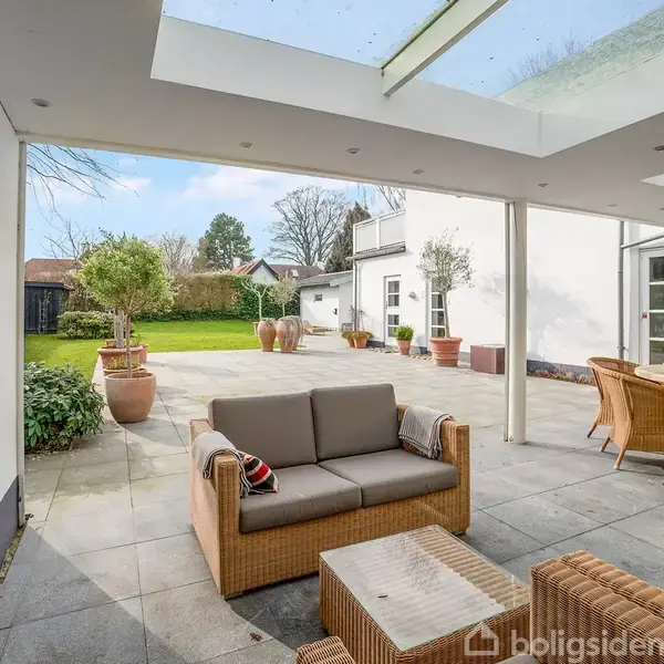 Sofagruppe står under en overdækket terrasse med glasloft, omgivet af en stor, grøn have med krukker og træer. Terrassebord med stole er også til stede.