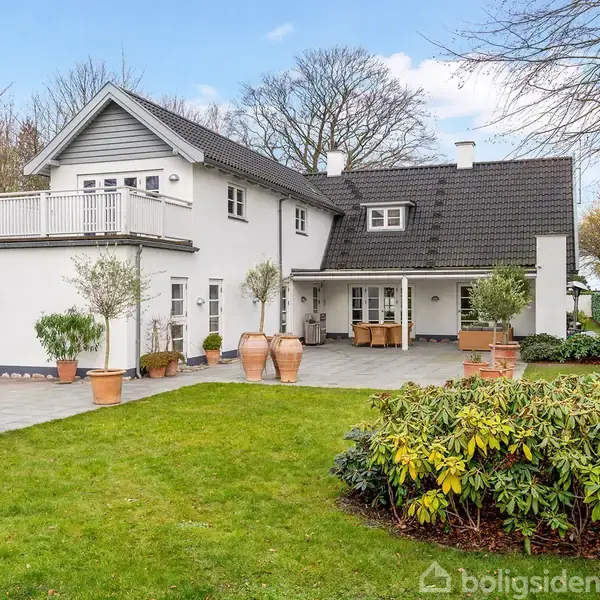 Hvid villa med sort tag står stille i en velplejet have med græs og buske. Et stort terrasseområde med krukker og træer giver en hyggelig udendørs atmosfære.