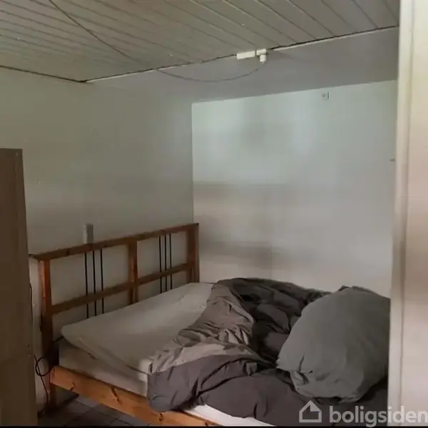 En seng ligger uopredt med mørkt sengetøj i et sparsomt møbleret værelse med hvidmalede vægge og træloft. På et skab står en blå flaske.
