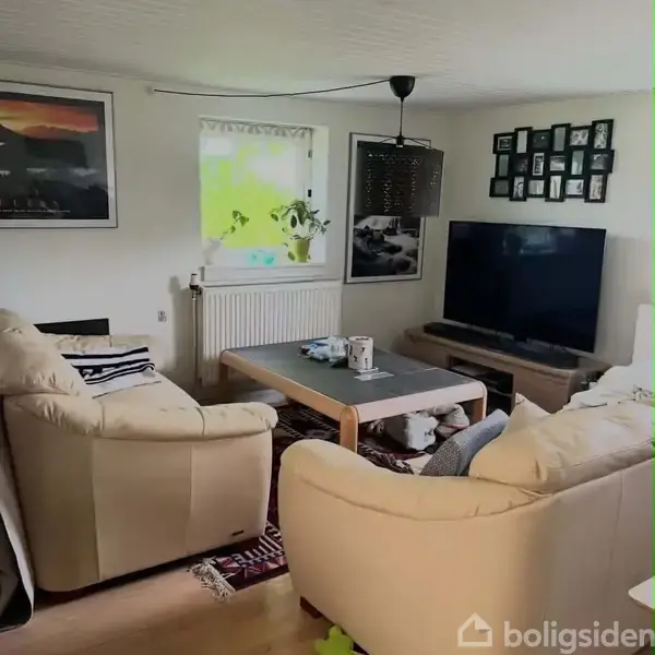 To beige sofaer og et sofabord står i en stue med en stor fladskærm på et TV-bord. Væggen har billeder og en lyskilde hænger fra loftet.