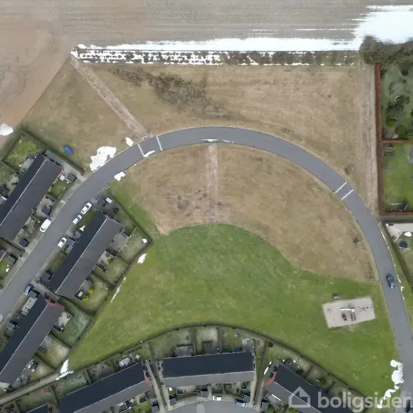 Et boligområde set ovenfra. Huse ligger langs en buet vej. Græsarealer og indkørsler omgiver bygningerne. Der er ingen tekst til stede.