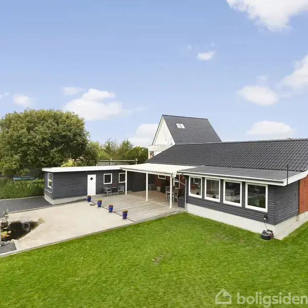En moderne etplans villa med fladt tag og store vinduer ligger i en velholdt have med en græsplæne og lille fiskedam. Villaen er omgivet af hække og andre grønne områder.