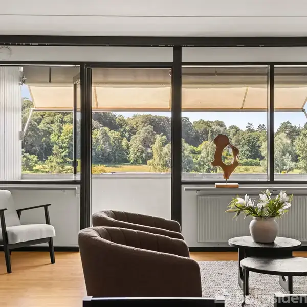 Stue med moderne møblering med stol, sofa og bord med blomster. Store vinduer giver udsigt til grønne træer og parklandskab udenfor. Farverne er lyse og indbydende.