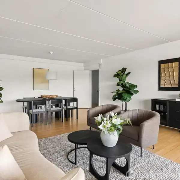 En stue med beige sofa og to grå stole, placeret omkring en sort rund bord med blomster, i et lyst rum med spisebord og planter.