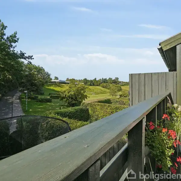 En træterrasse med blomster og udsigt over en grøn have med træer og buske. En sti snor sig gennem landskabet under en blå himmel.
