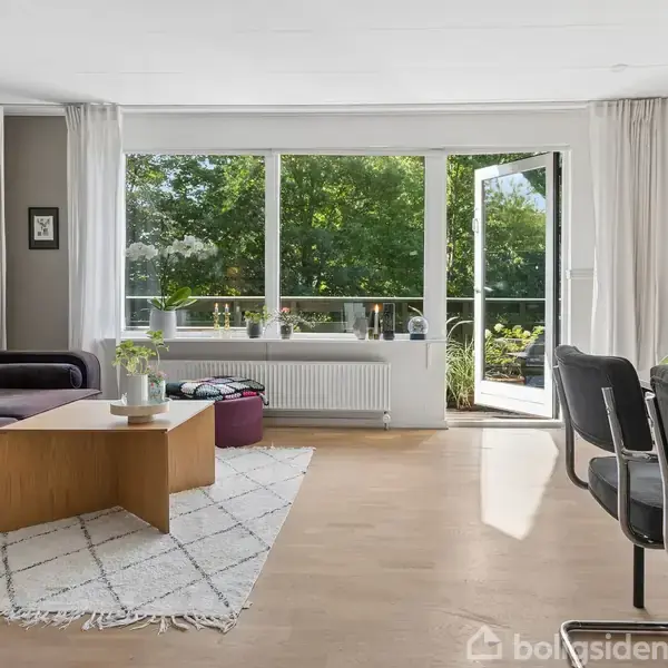 Stue med en mørk sofa og træbord, sollys strømmer ind gennem store vinduer, som åbner ud til en balkon med udsigt til grønne træer. Blødt tæppe dækker gulvet.