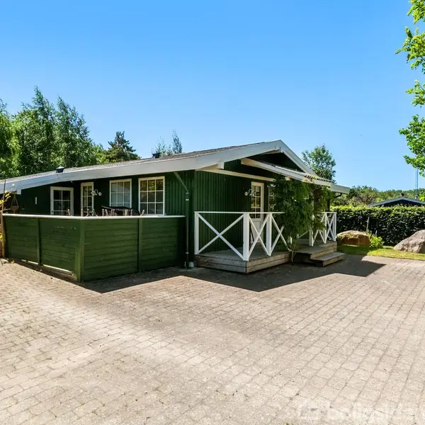 Et grønt træhus står på en brolagt gårdsplads omgivet af frodige træer og buske. Foran huset er der en veranda med hvide gelændere. Himmel er klar og blå.