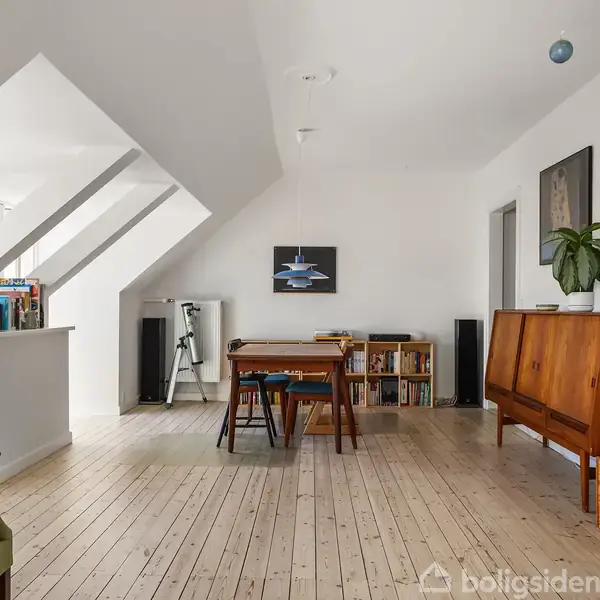 Et spisebord med stole står centralt i et lyst rum med trægulv. Ved siden af er en reol fyldt med bøger. Væggene er hvide, der hænger en lampe og et maleri.