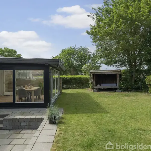 En terrasse med glasindrammet veranda, der vender mod en have med græsplæne. I baggrunden står et lille træhus omgivet af grønne buske og træer, under en klar himmel.