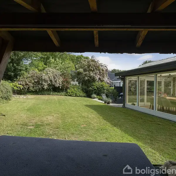 Altan udsigt over en grøn have med velplejet græsplæne og buske. Til højre ses en glaskarnap på huset, der åbner op mod det lyse udendørsområde.