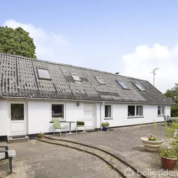 Hvidt hus med gråt tag har flere vinduer og en hoveddør. Står i gårdhave med flisebelægning og blomsterkrukker. Omkringliggende grønne planter og en klar himmel.