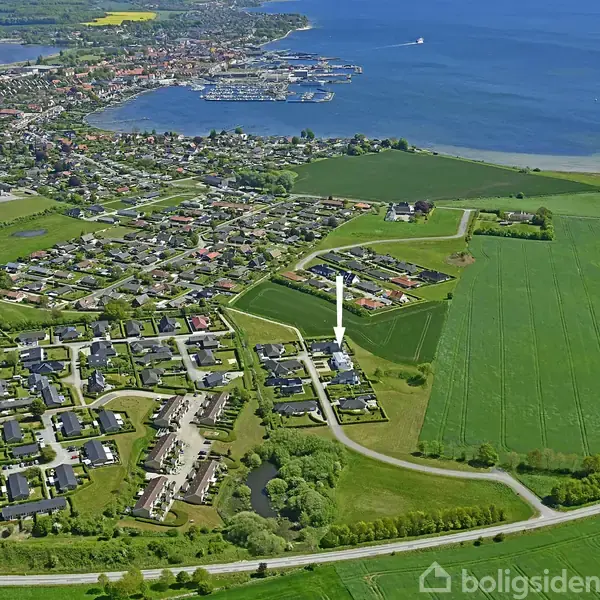 Et boligområde strækker sig ud langs kysten med et havnen i baggrunden. En pil peger mod et specifikt hus i nabolaget, omgivet af grønne marker og veje.